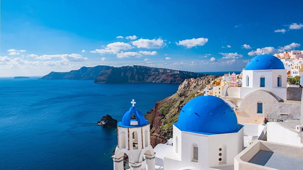 View of Santorini’s blue-domed churches overlooking the Aegean Sea on a sunny day—iconic Greek island scenery for family beach travel in Greece.