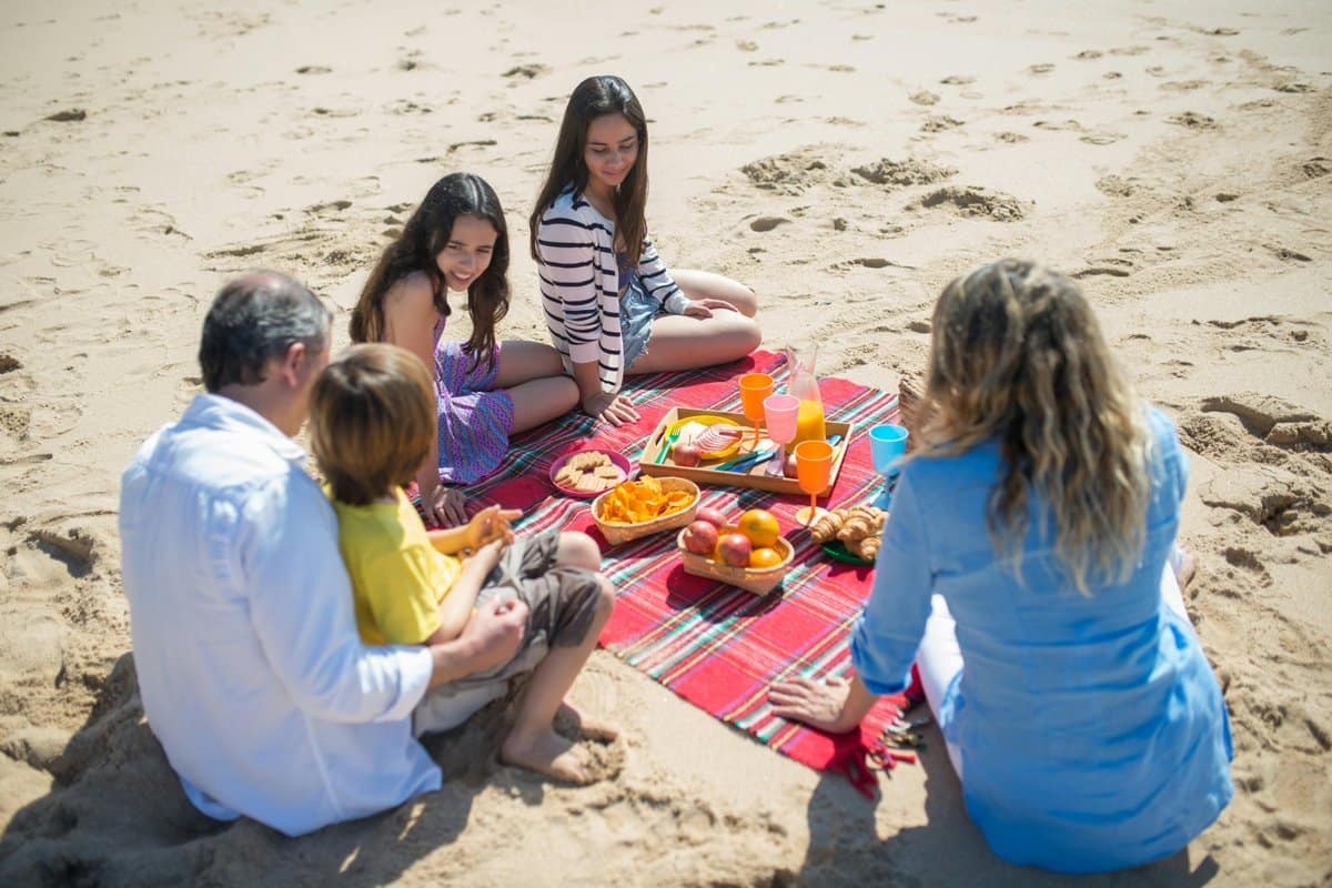 family-beach-blanket-sand-free-travel-1200x800 | Go With The Family Family sitting together on a beach blanket during a seaside picnic — a great example of a sand-free or easy-shake beach blanket for family trips.