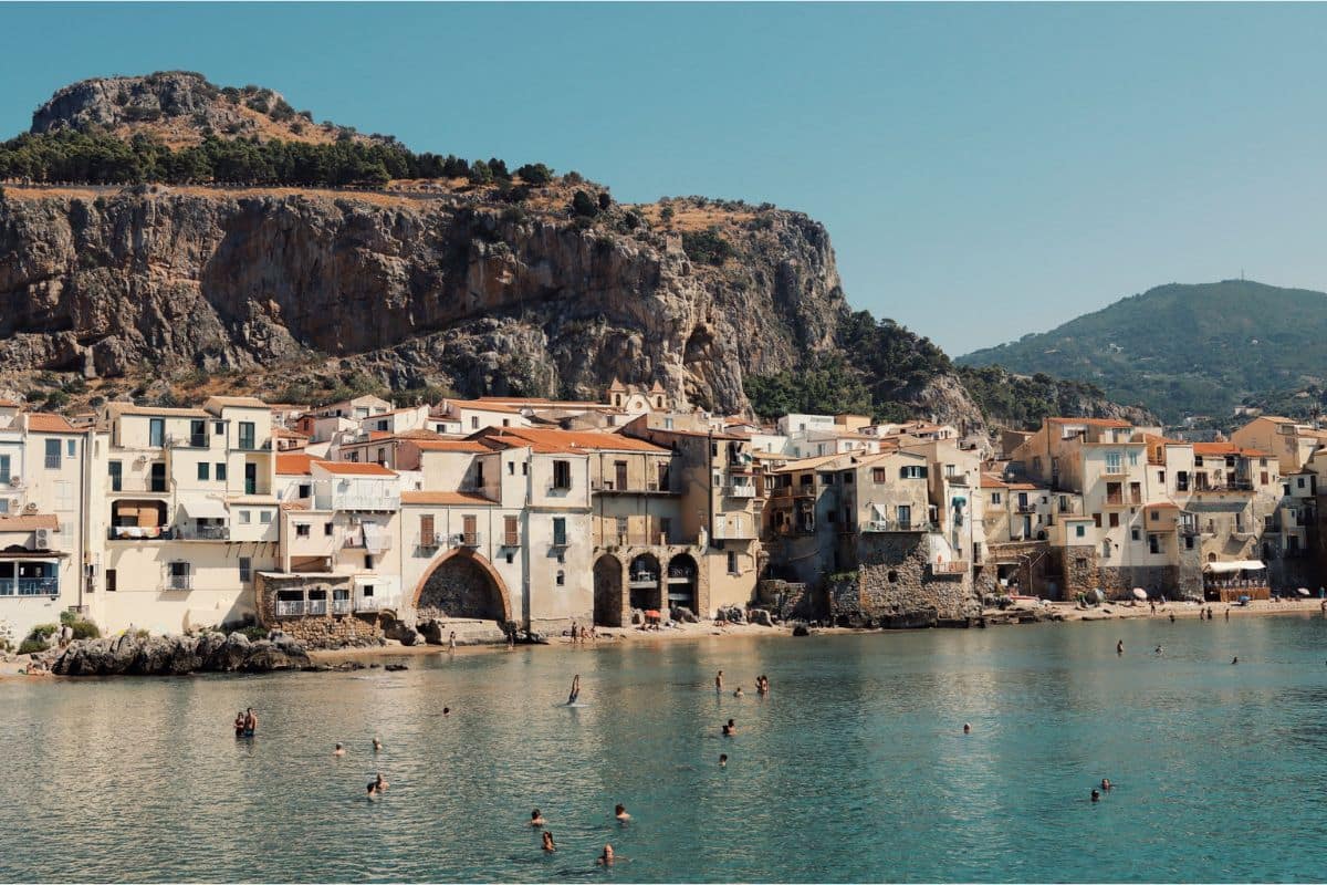 Cefalù Old Town, Sicily | Go With The Family Historic waterfront of Cefalù in Sicily with sandy beach, colorful old town buildings, and crystal-clear Mediterranean water.
