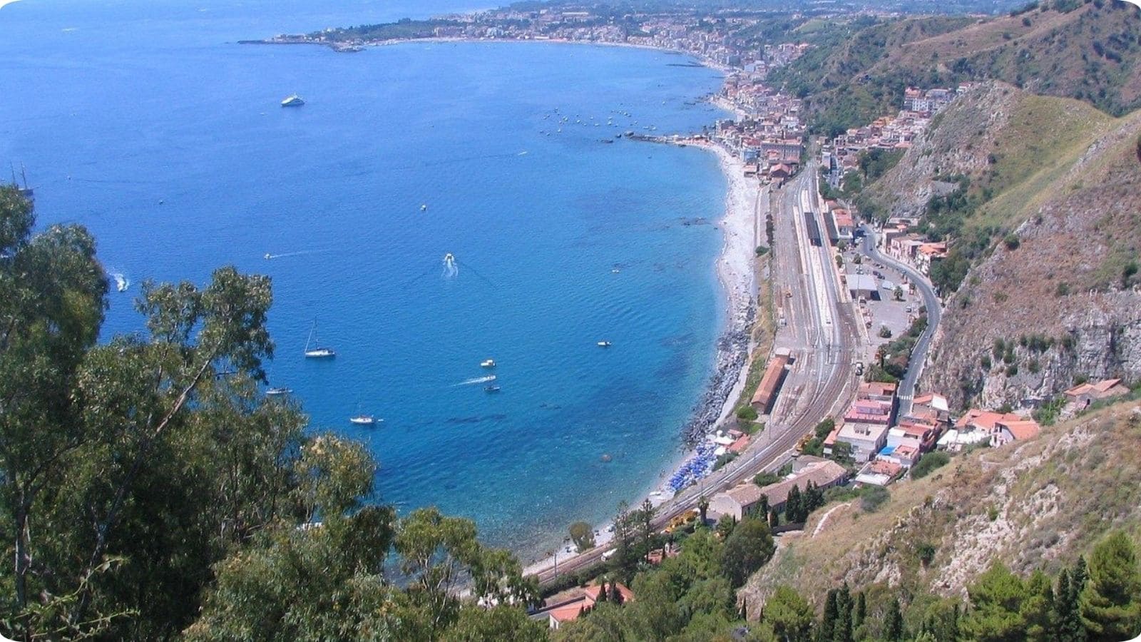 Taormina Coastline, Sicily | Go With The Family Panoramic view of the Taormina coastline in Sicily with turquoise Mediterranean water, beaches, and dramatic cliffs.
