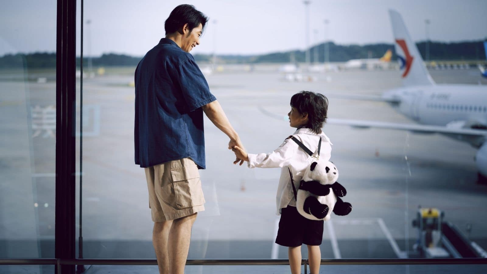 Family Travel at the Airport | Go With The Family Parent and child looking out at an airplane through the airport terminal window before a family flight.